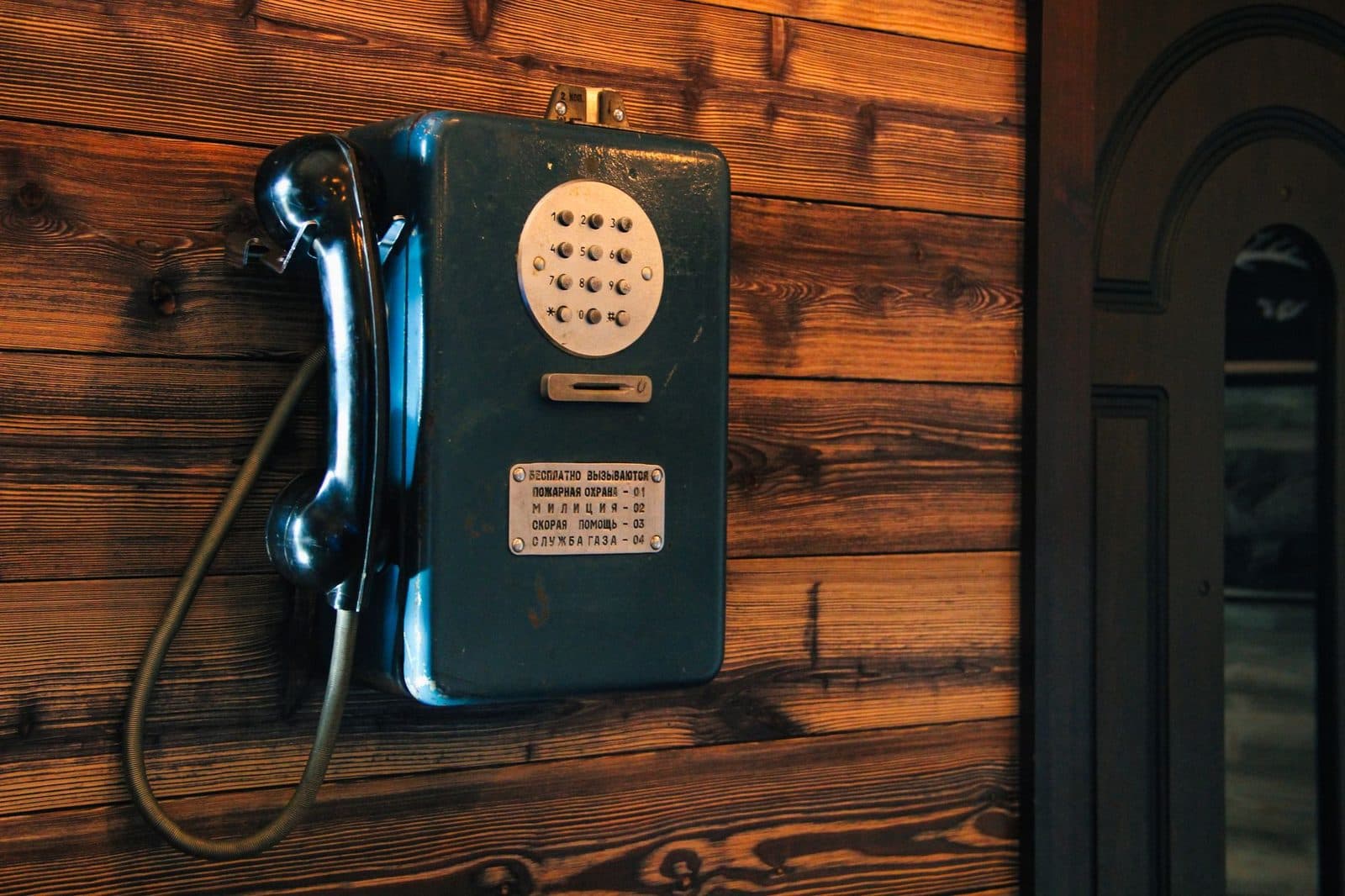 Teléfono mural analógico azul con auricular fijado a una pared de madera, junto a una placa con números de emergencia en cirílico
