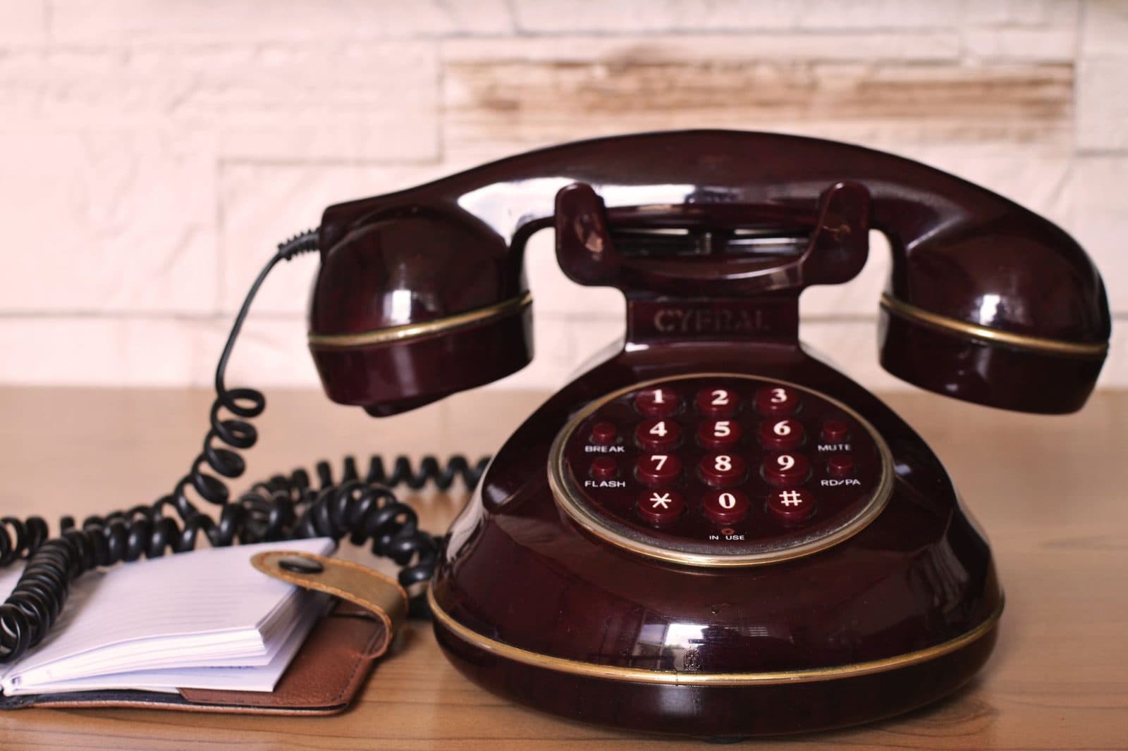 Vintage burgundy landline telephone with keypad and coiled cord on a wooden table, next to a notebook