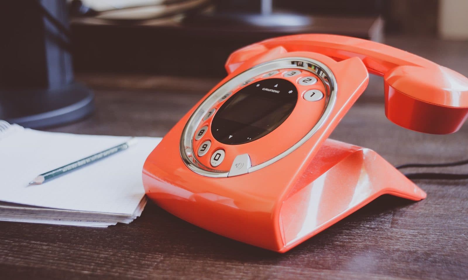 Telefono fisso retrò arancione appoggiato su una scrivania di legno, accanto a un blocco per appunti e a una matita