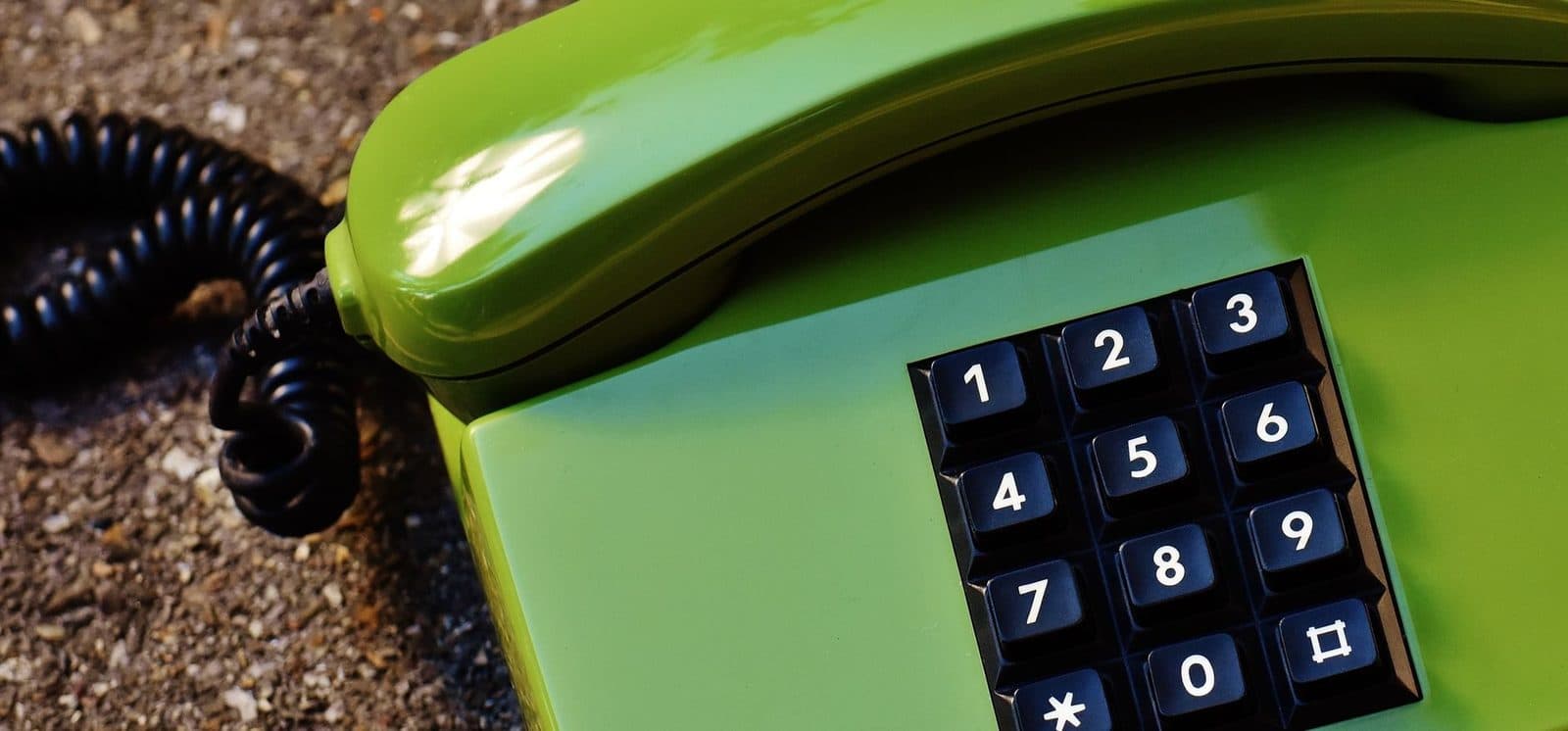Telefono fisso verde con cornetta a filo e tastiera con tasti neri, appoggiato sulla ghiaia