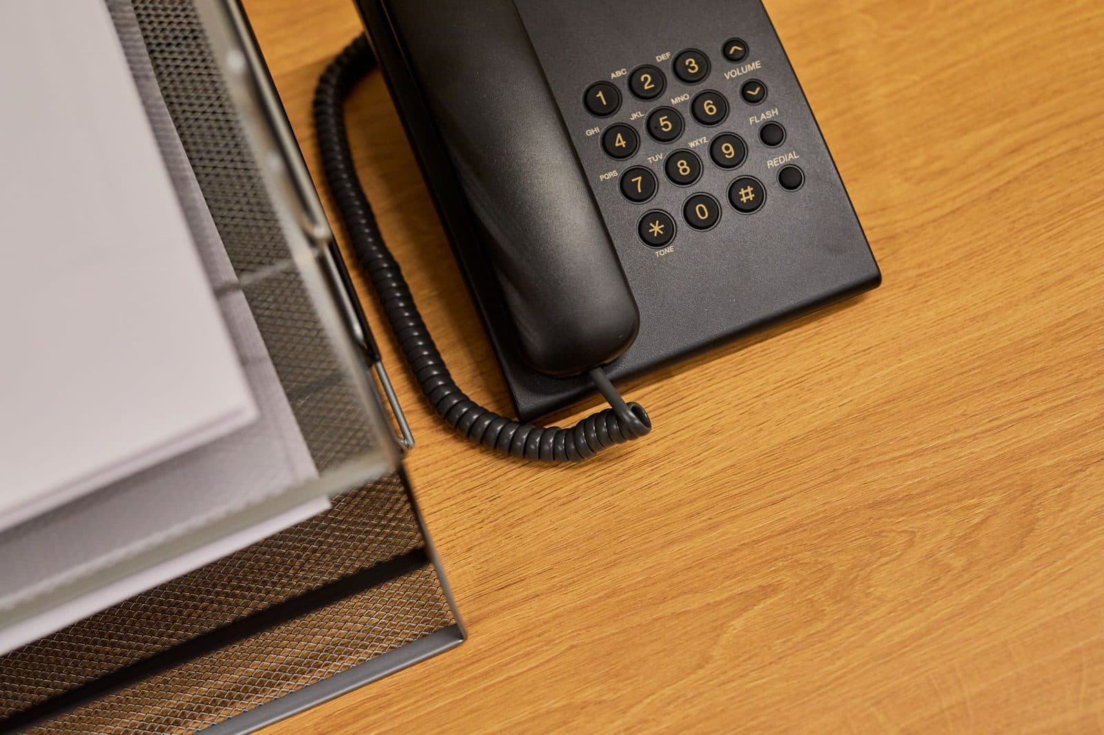 Black push-button landline telephone with handset and coiled cord, on a wooden desk next to signature folders