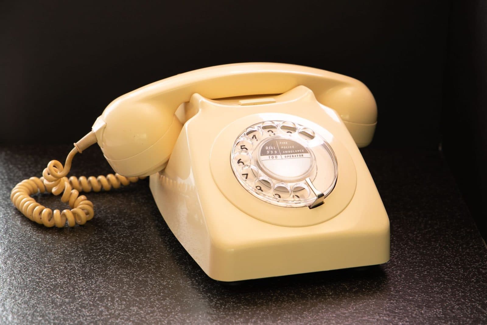 Vintage ivory rotary-dial landline telephone with handset and coiled cord, sitting on a dark cabinet