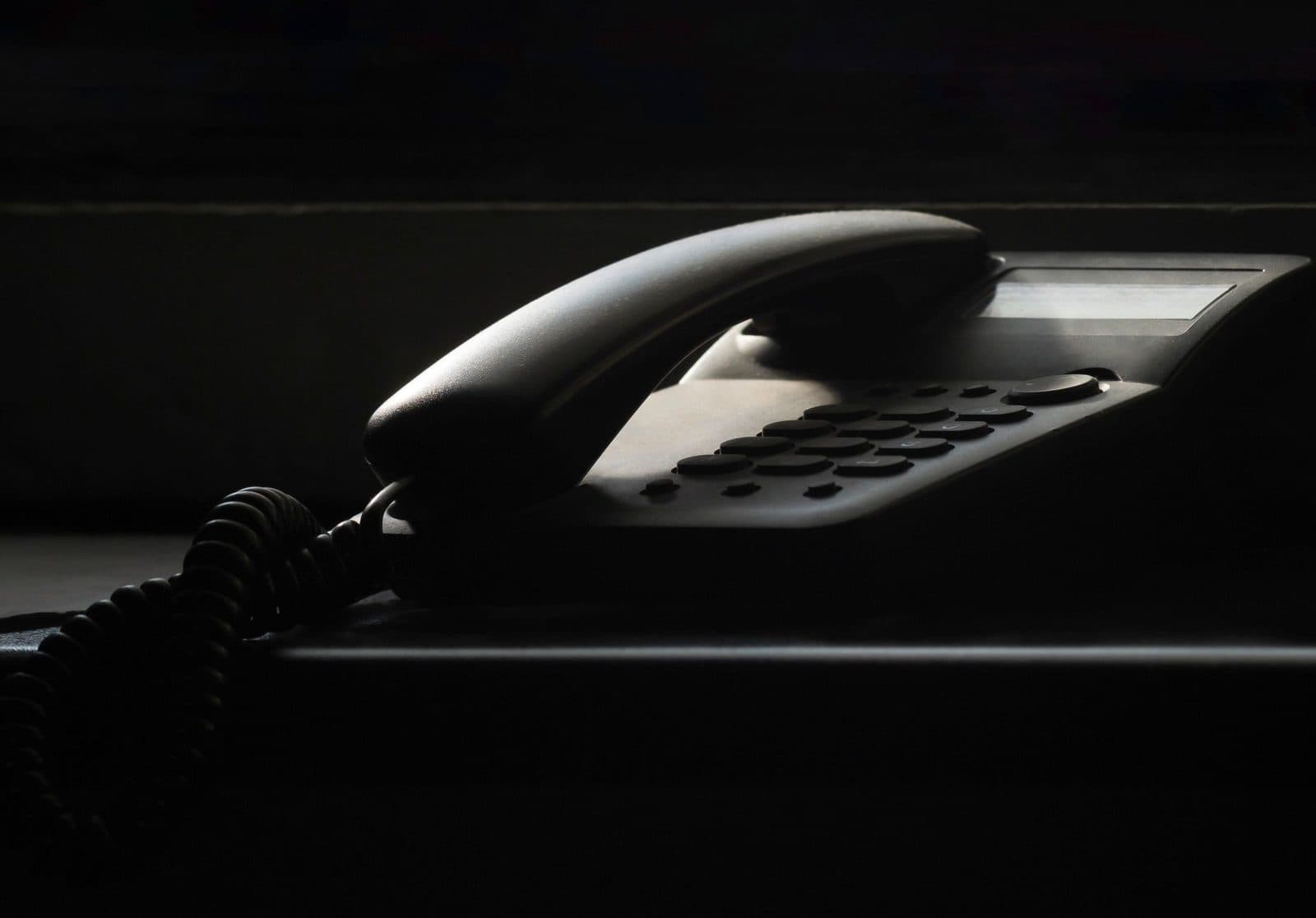 Schwarzes Festnetztelefon mit Hörer und Tastenfeld auf einem Möbelstück, in einem dunklen, schwach beleuchteten Raum