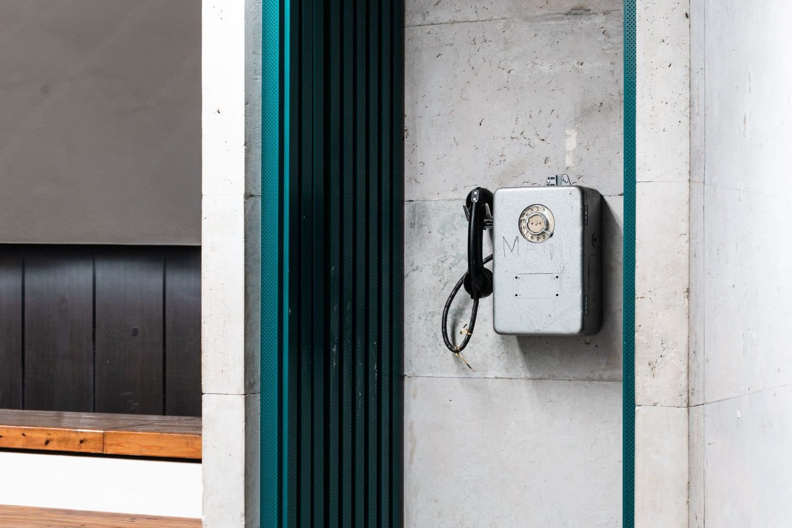 Old grey wall-mounted rotary dial telephone with a black handset hanging, fixed to a concrete wall