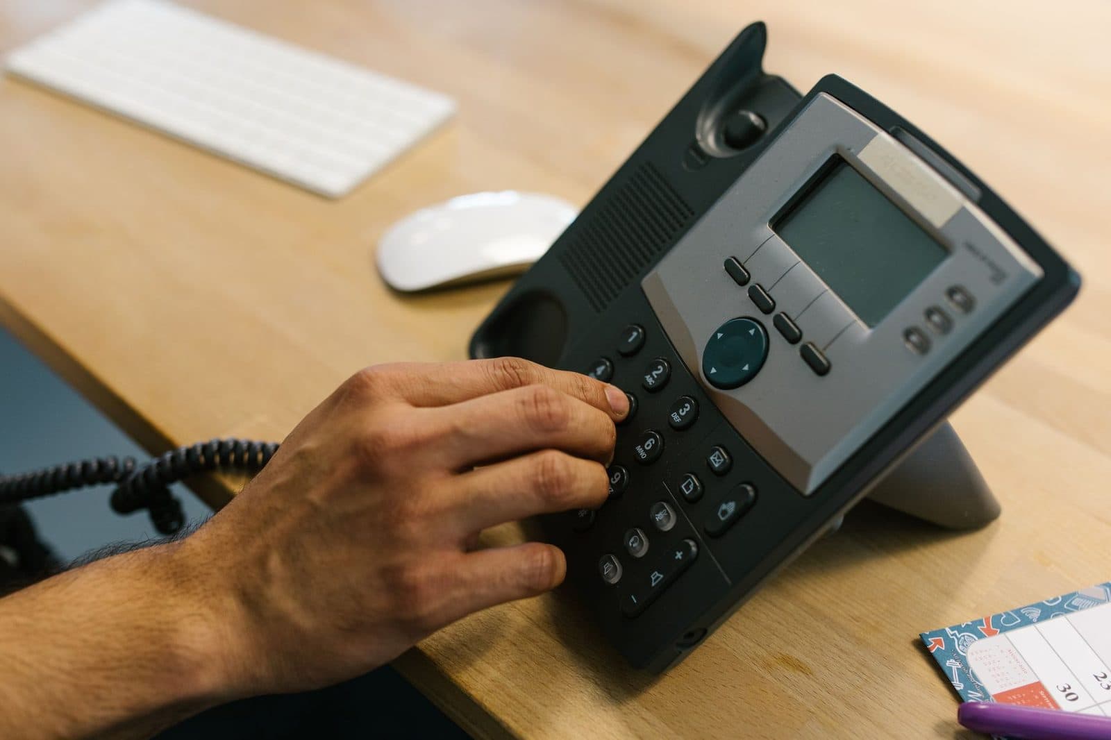 Mano che compone un numero su un telefono fisso da ufficio appoggiato a un tavolo, accanto a una tastiera e a un mouse