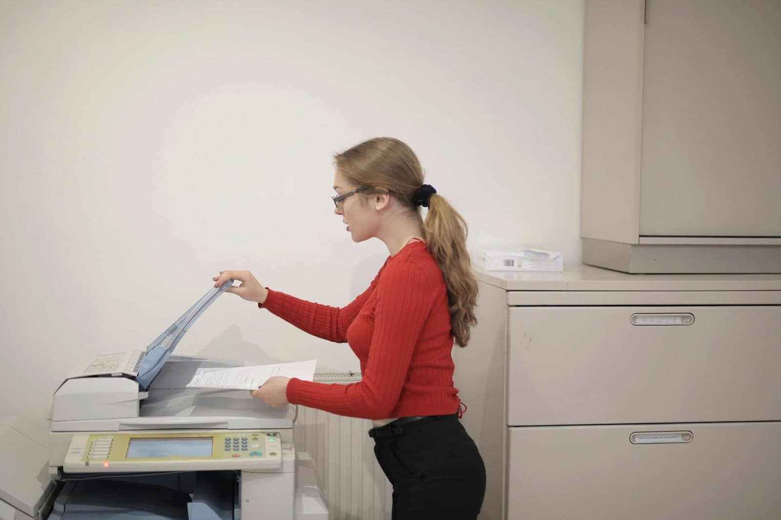 Junge Frau im roten Pullover legt in einem Büro ein Blatt in ein Multifunktionsgerät mit Faxfunktion ein
