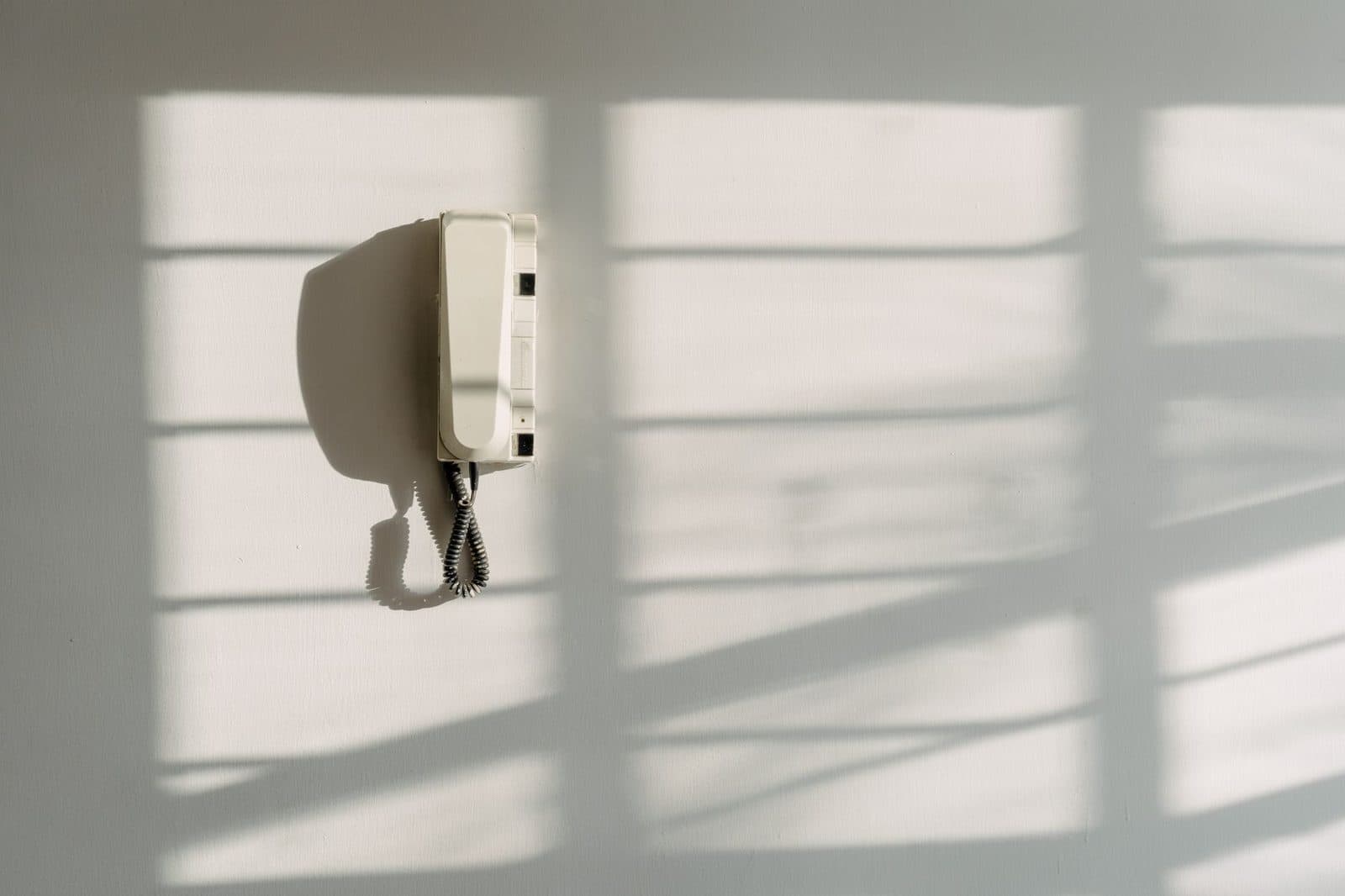Weißes analoges Wandtelefon mit Spiralkabel an einer hellen Wand, überzogen vom Schatten einer Jalousie