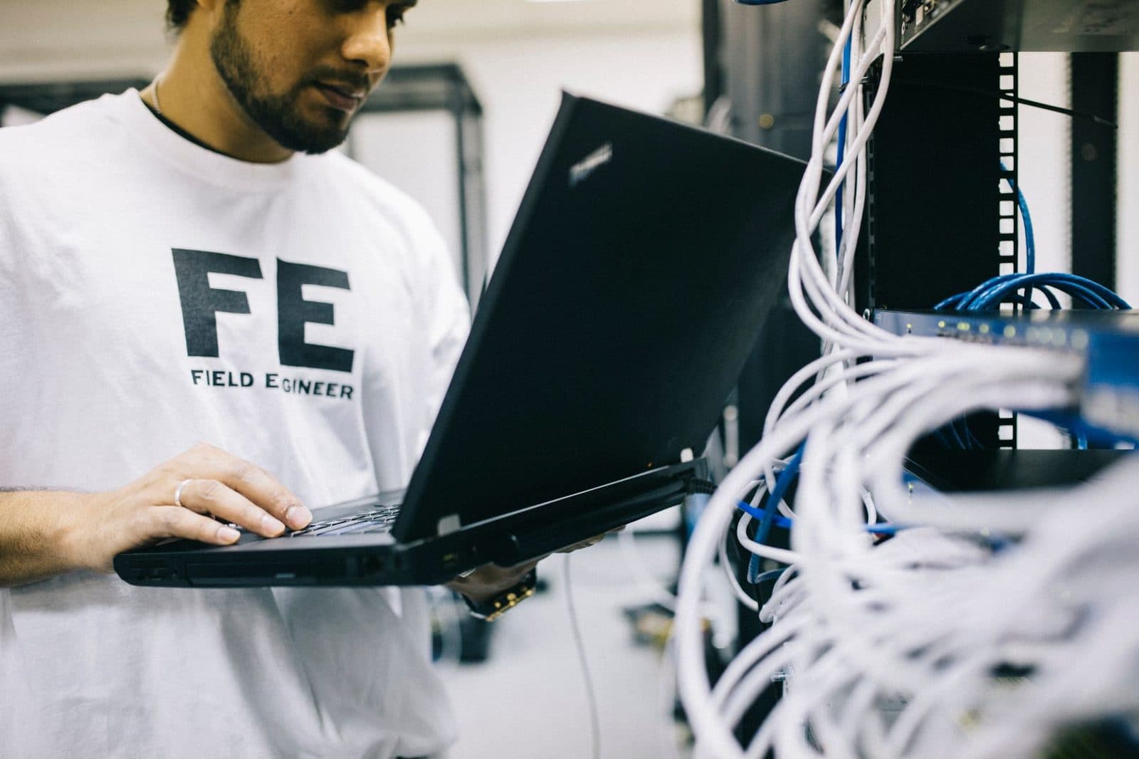 Technician in a "Field Engineer" T-shirt with a laptop in front of a patch panel rack and network cables
