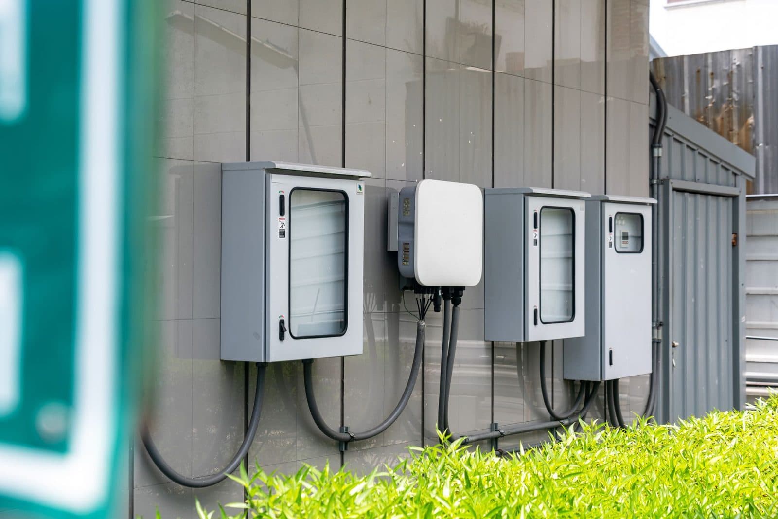 Grey telecommunications cabinets mounted on an exterior wall, exposed cables and a green hedge