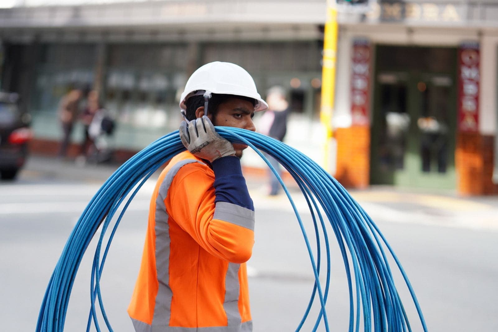 Tecnico con gilet arancione e casco bianco che trasporta una matassa di cavi blu in una strada