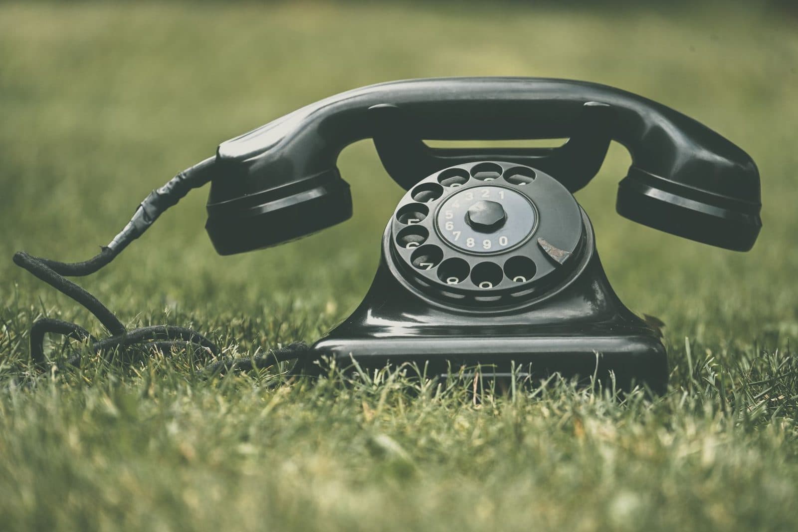 Black rotary dial landline telephone, handset off the hook, lying in the grass