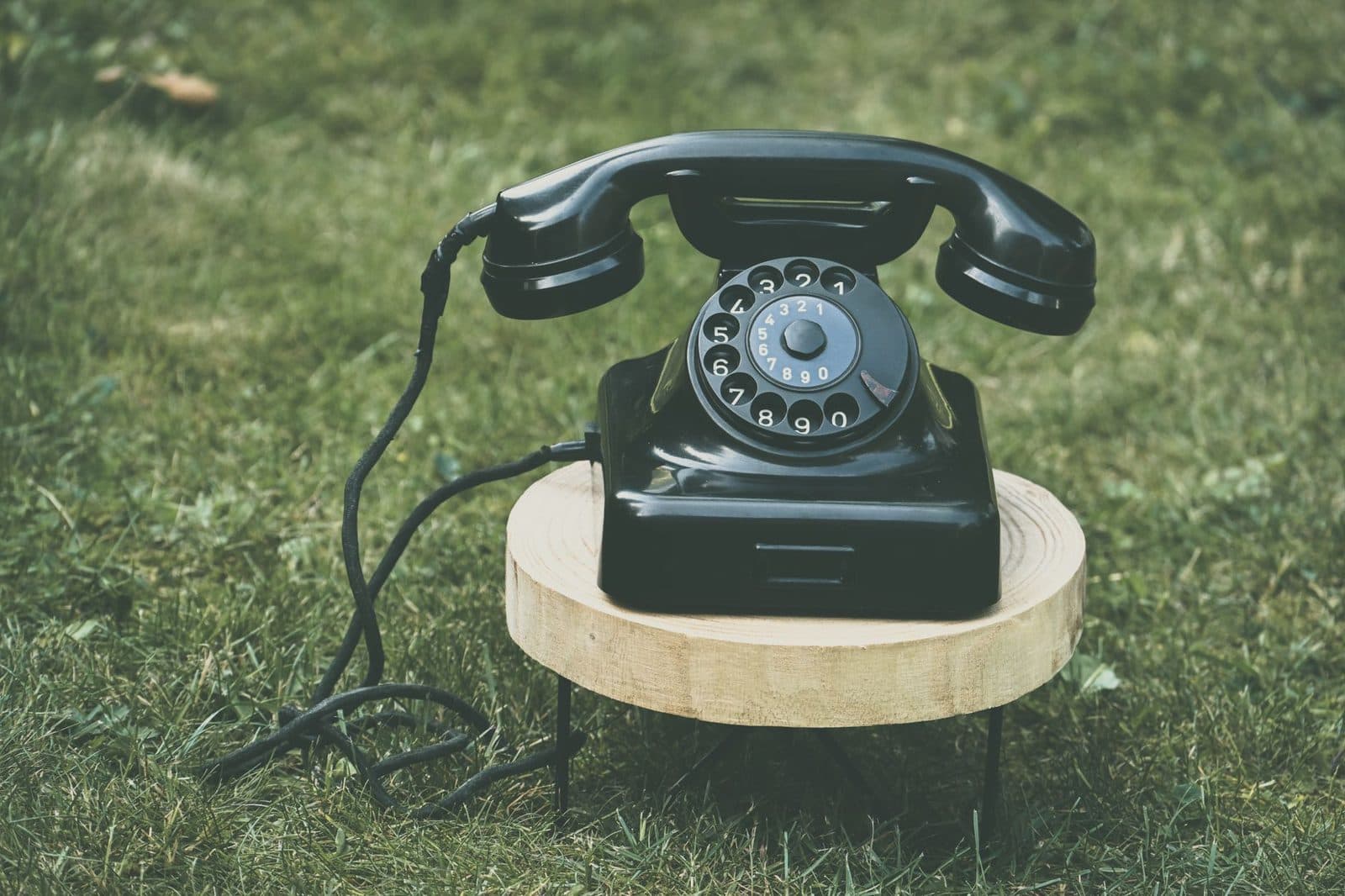 Black rotary dial telephone sitting on a wooden slice in the grass, cable unspooled