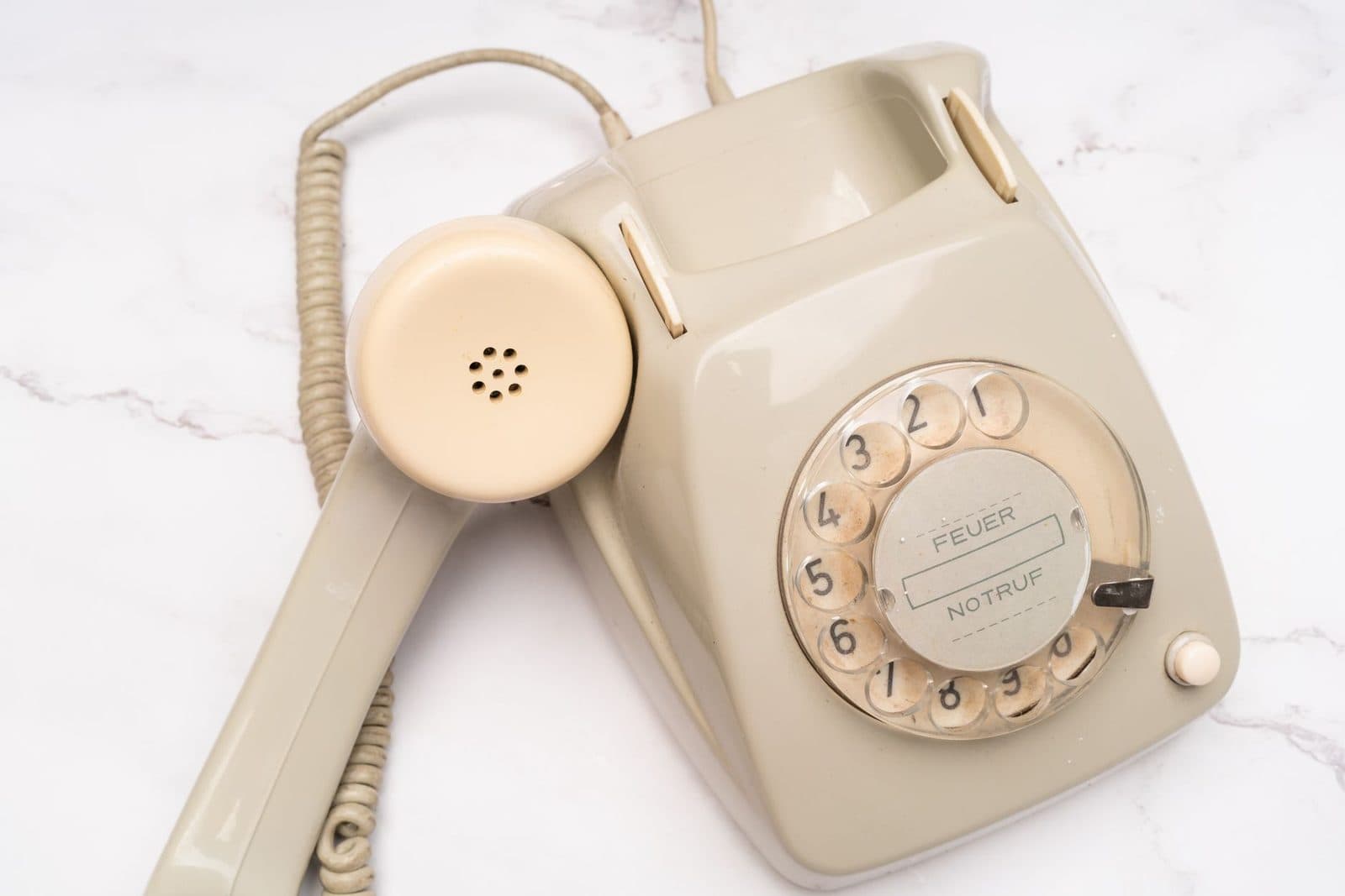 Altes beigefarbenes Festnetztelefon mit Wählscheibe, abgenommener Hörer, auf einer weißen Marmorplatte