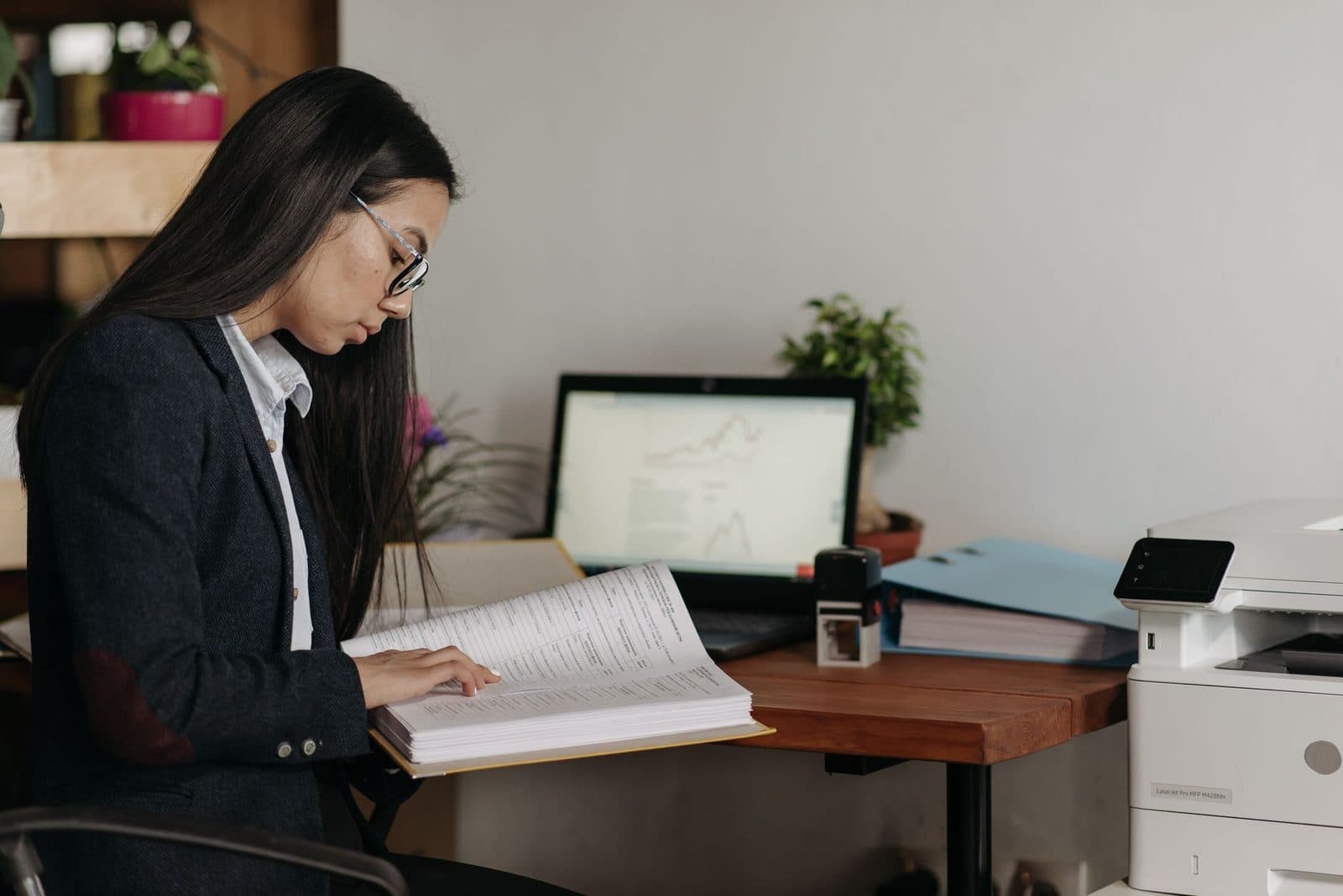 Femme en veste consultant un dossier papier dans un bureau, près d'un ordinateur portable et d'une imprimante multifonction