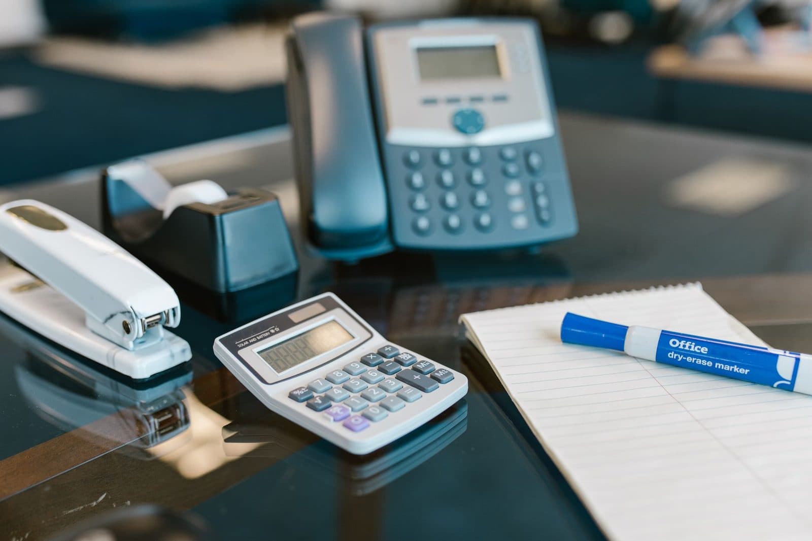 Bürotelefon auf einem Glastisch, daneben Taschenrechner, Hefter, Notizblock und Marker