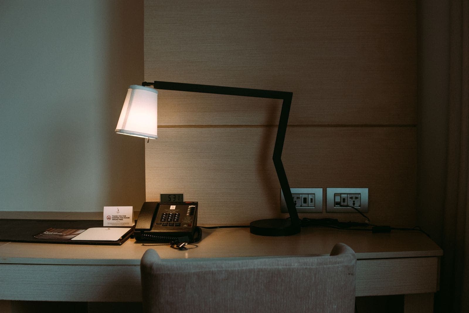 Telefono fisso con filo appoggiato su una scrivania in legno illuminata da una lampada, vicino a prese a muro